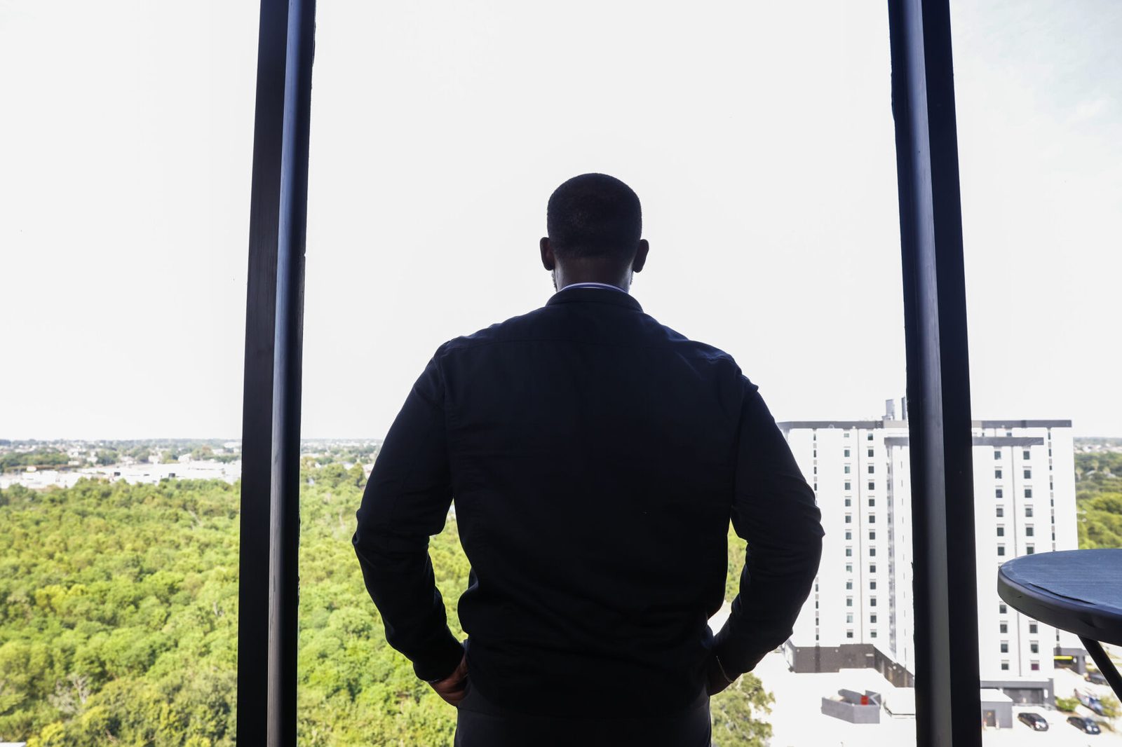 Oel Scott, seen from behind, looks out a tall window over the treetops and buildings of New Orleans East.
