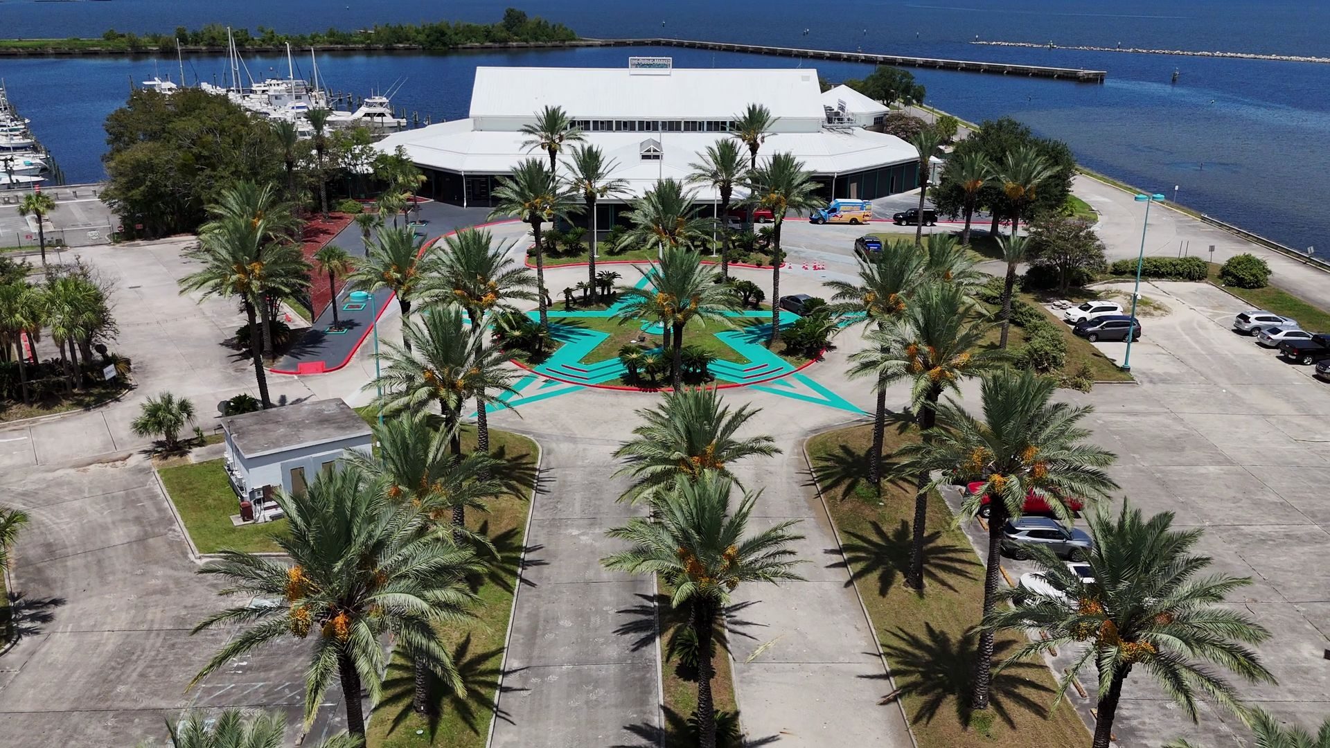 Aerial view down the palm-lined entry drive to the market building, with the marina and Lake Pontchartrain behind.